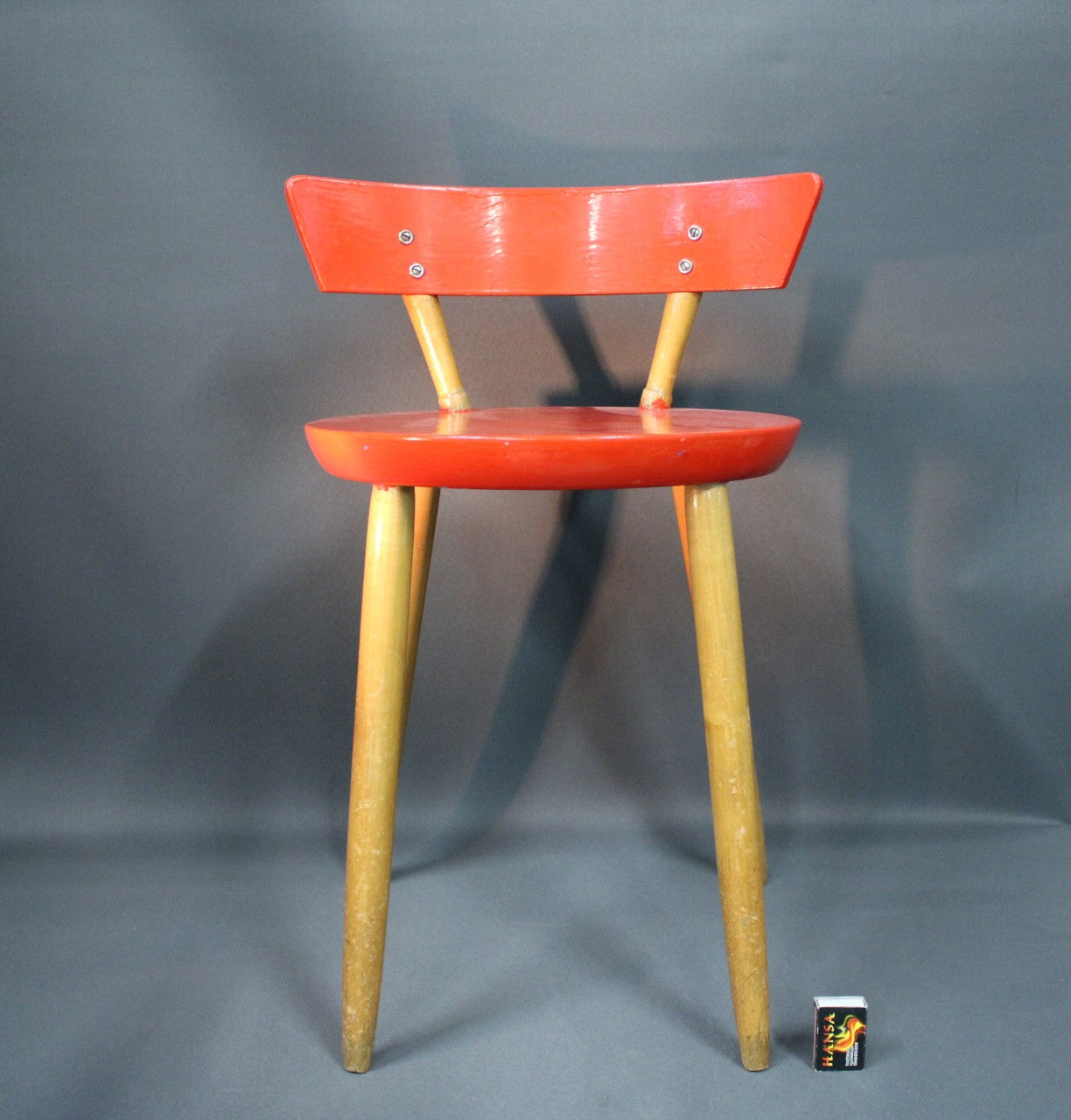 Puinen 1950-luvun tuoli, punainen istuin ja selkänoja,tulitikkurasia hahmottamassa kokoa